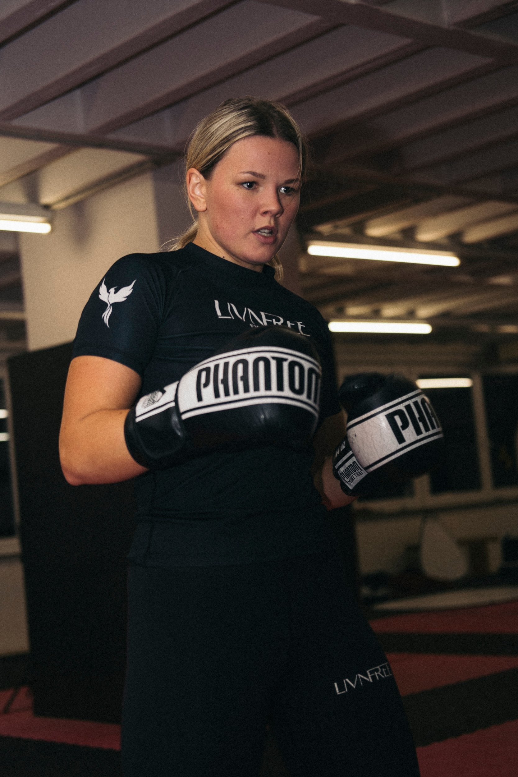 Frau in Sportkleidung mit LIVNFREE-Logo und schwarzen 'Phantom'-Boxhandschuhen trainiert konzentriert im Fitnessstudio, Nahaufnahme mit intensivem Blick