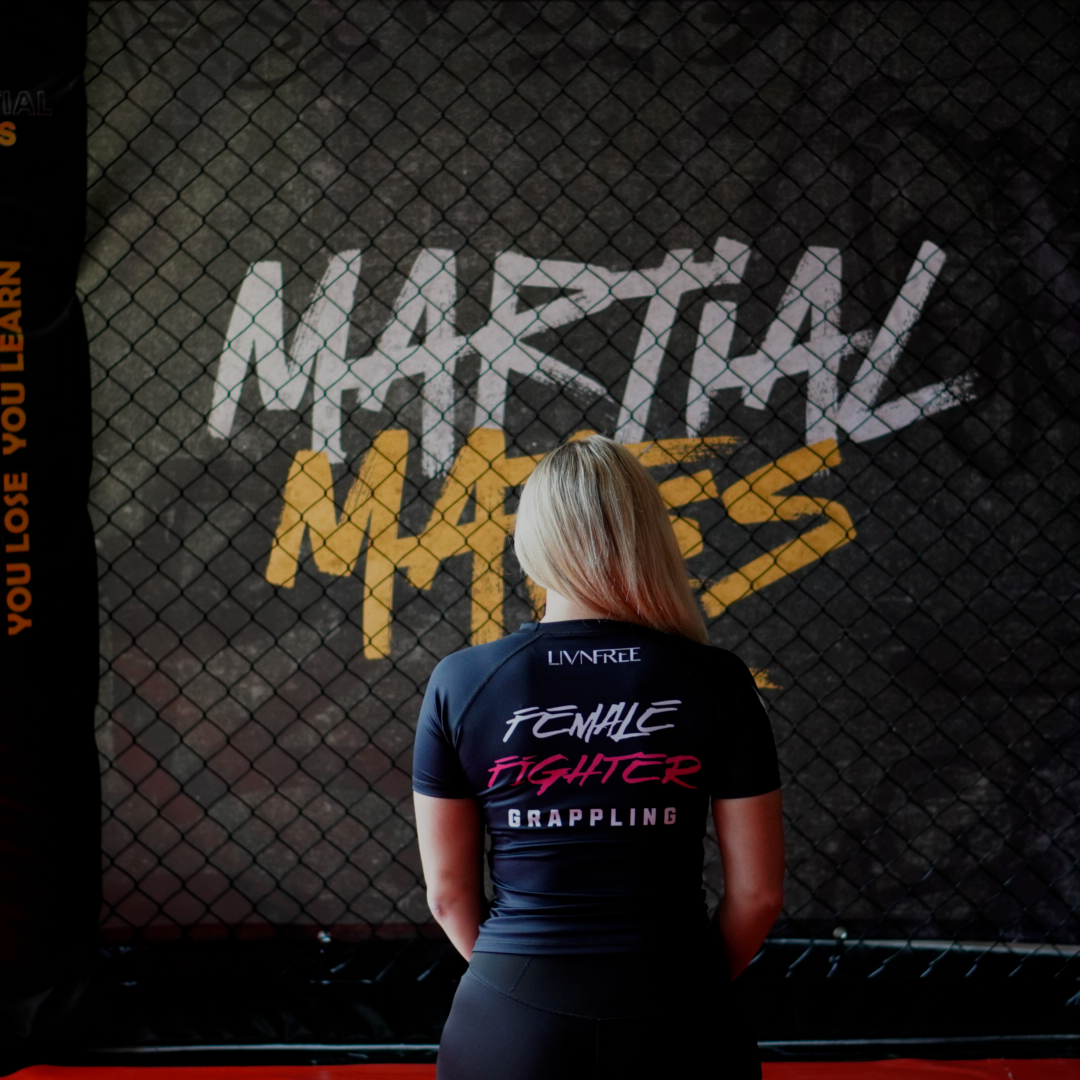 Weibliche Kämpferin in Rash Guard mit der Beschriftung Female Fighter Grappling vor einem Martial Mates Hintergrund