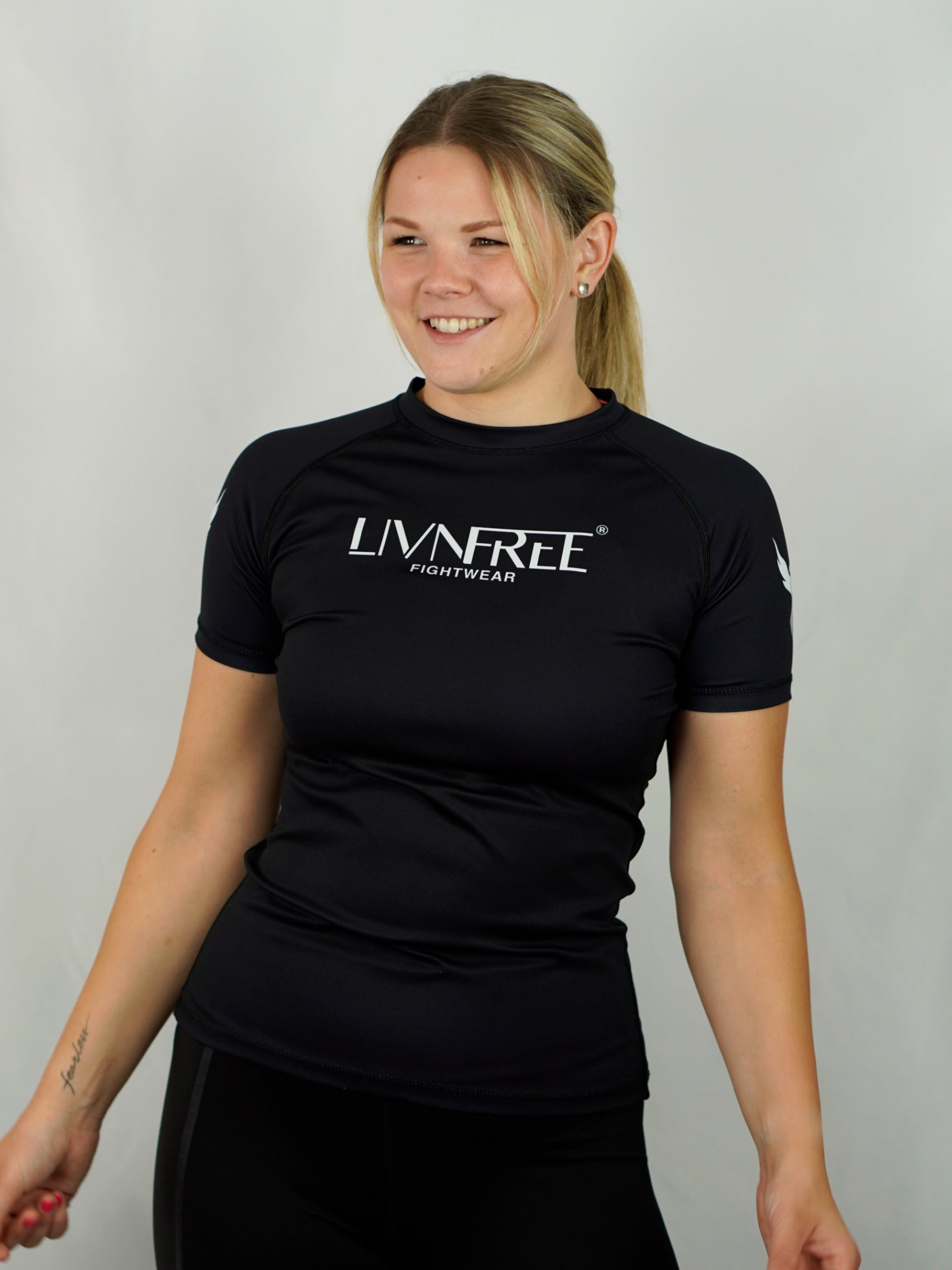 Frau in 'LivnFree'-Sportshirt mit Zopf lächelt und blickt zur Seite, entspannte Körperhaltung, vor hellem Hintergrund